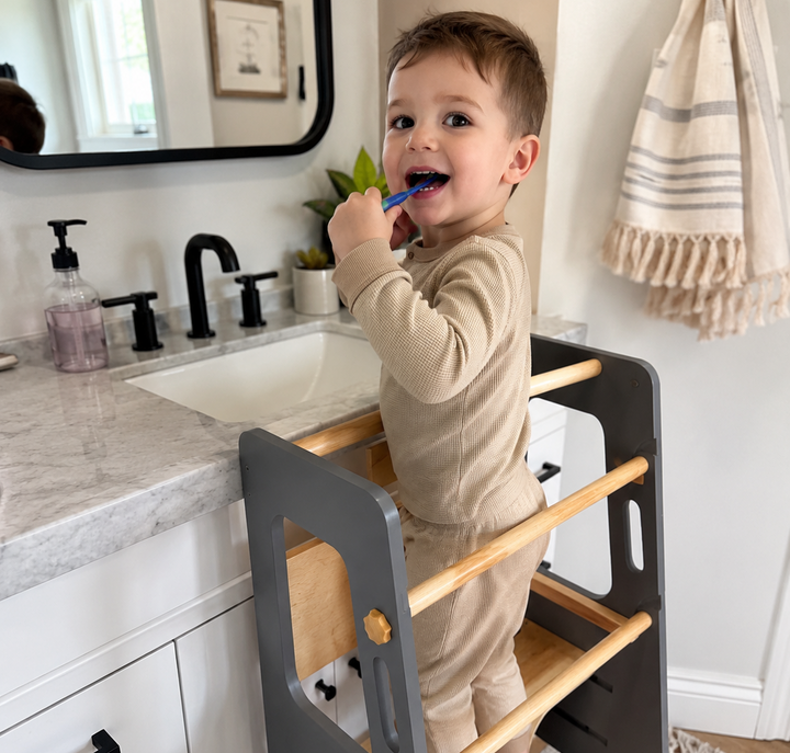 Toddler brushing teeth independently with a gray Montessori learning tower at the bathroom sink